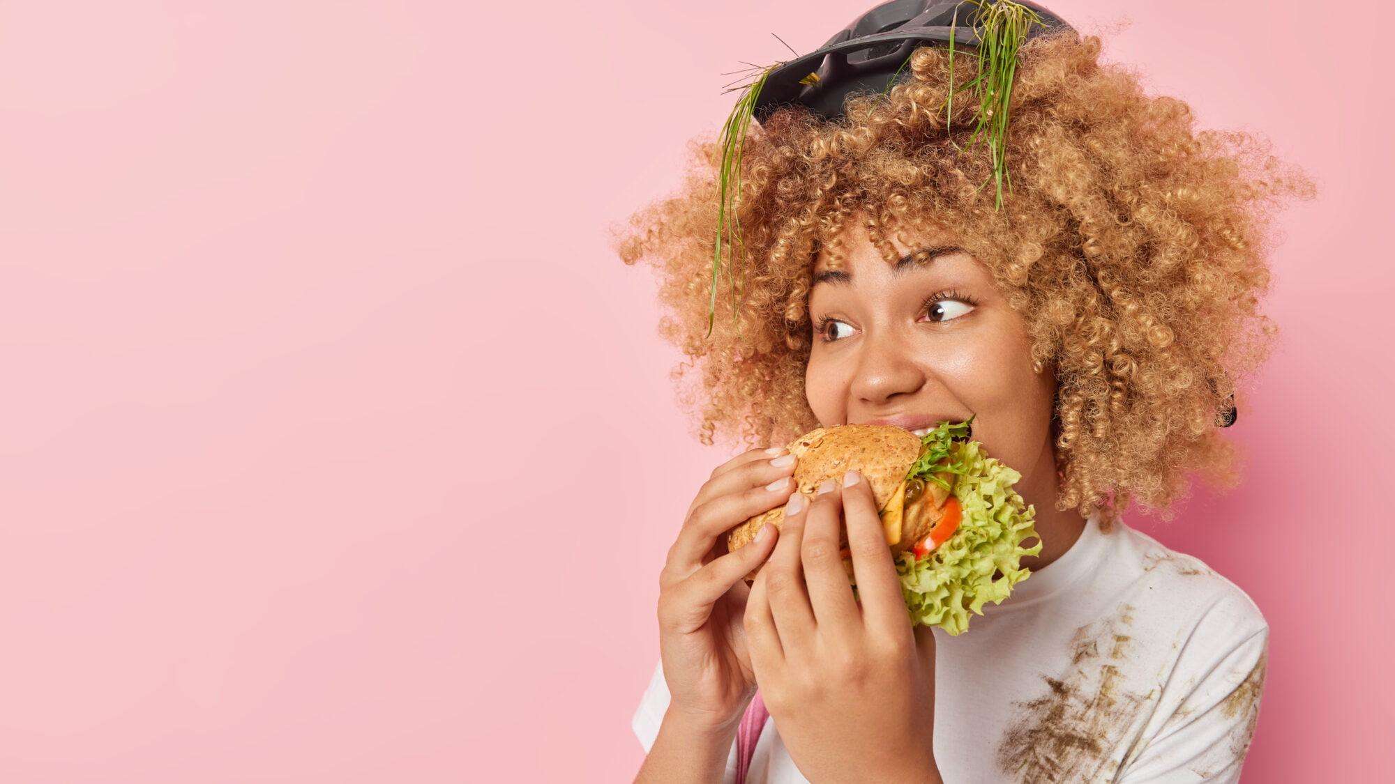 Photo of curly haired woman wears protective helmet for safe riding on bicycle eats delicious sandwi